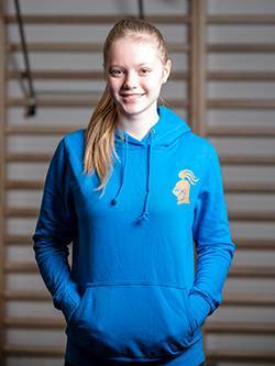 Kapuzenpulllover_Damen_vorne Ein sportliches Mädchen steht vor einer Sprossenwand in einem Fitnessstudio und trägt einen blauen Damen-Kapuzenpullover mit einem orangenen fitterritter Logo.
