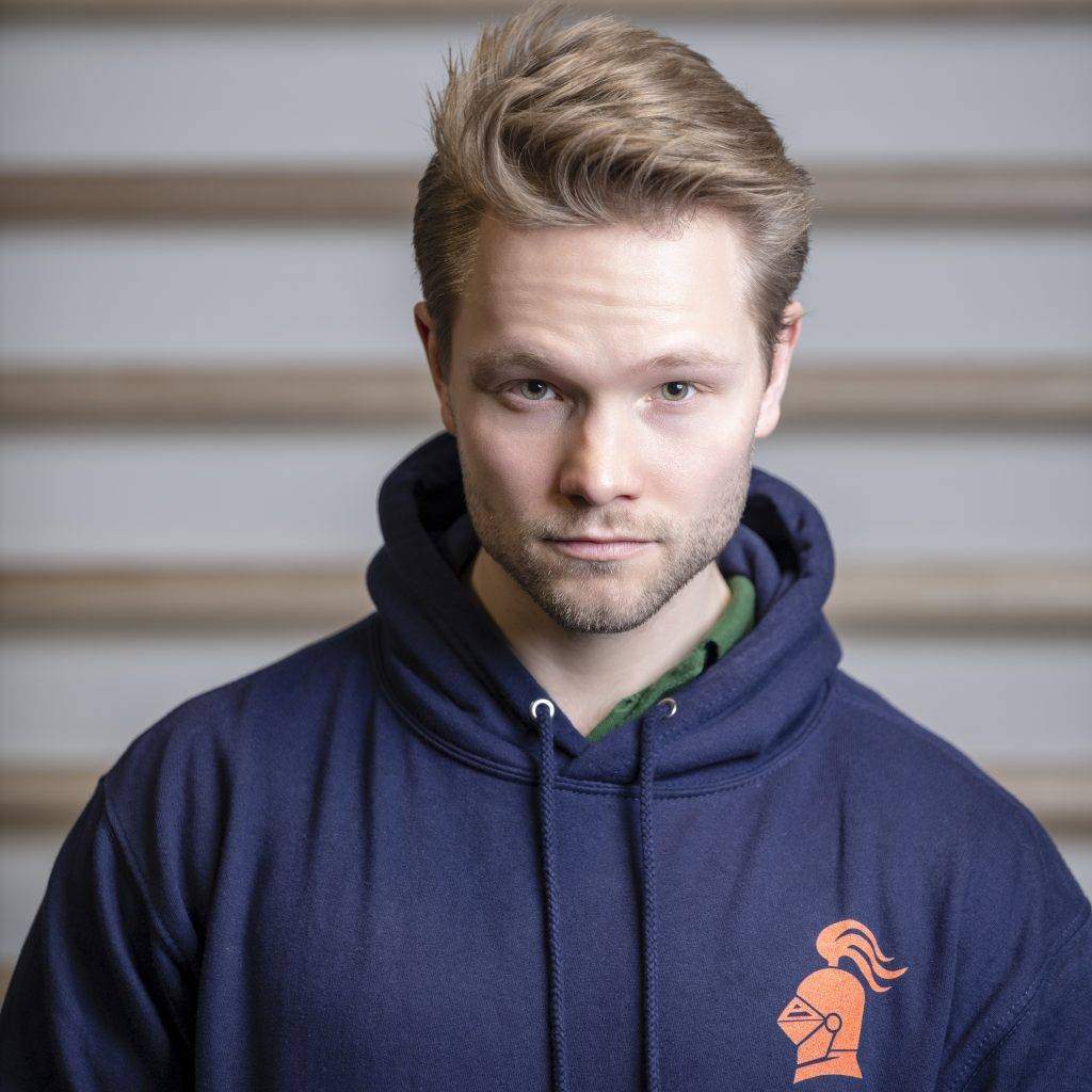 Shopfoto Männerpullover vorne Personal Trainer Jakob Ritter steht vor einer Sprossenwand und posiert mit einem dunkelblauen Herren-Kapuzenpullover, den ein orangenes fitterritter Logo ziert.