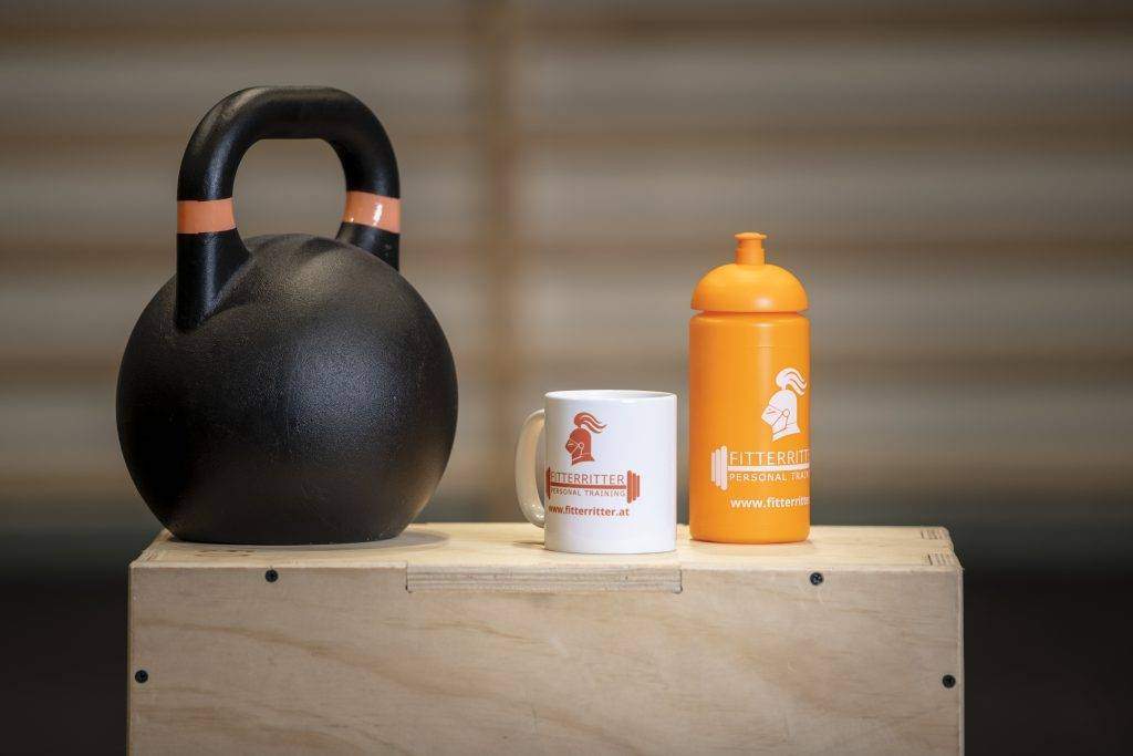 Shopfoto Trink dich fit Combo Auf einem Holztisch in einem Fitnesscenter befinden sich eine weiße fitterritter Tasse und eine orangene fitterritter Trinkflasche. Die Combo tritt unter dem Namen „Trink dich fit“ auf.